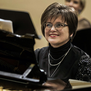 Oksana Rapita, a woman with short brown hair and glasses, sits at a piano and smiles.