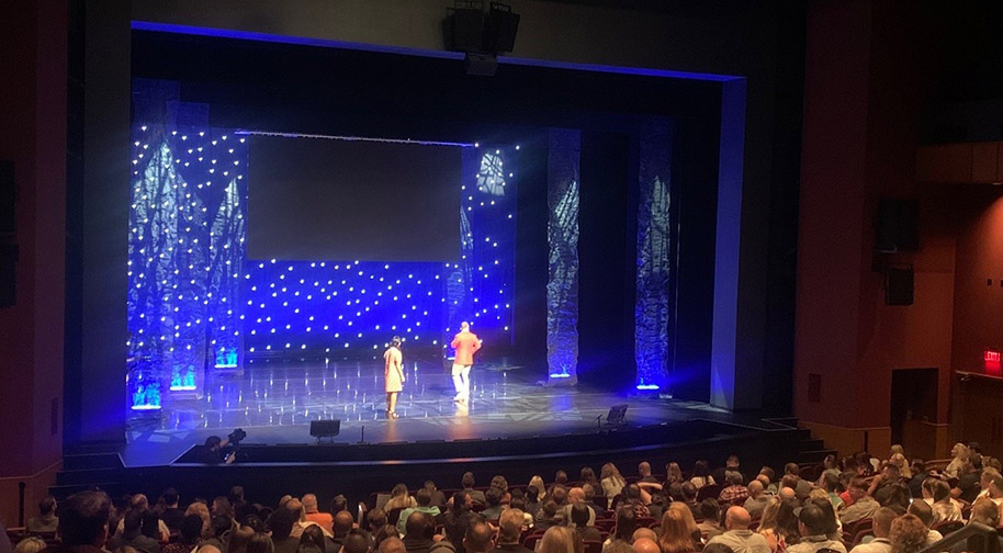 A corporate event in the Tarkington theater. Two people present on stage against a blue background dotted in white stars and patterns.