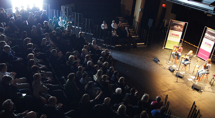 A daytime concert in the Studio Theater. Three acoustic guitarists on stools perform for a full audience. Curtains are parted at the windows to let daylight illuminate the stage.