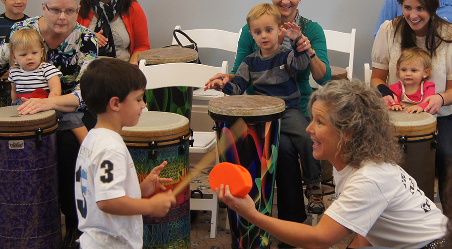 Instructor with curly gray hair and an excited expression holds out a percussion instrument toward a young child who hits it with a drumstick. In the background, other young children sit in the laps of their parents while playing hand drums.