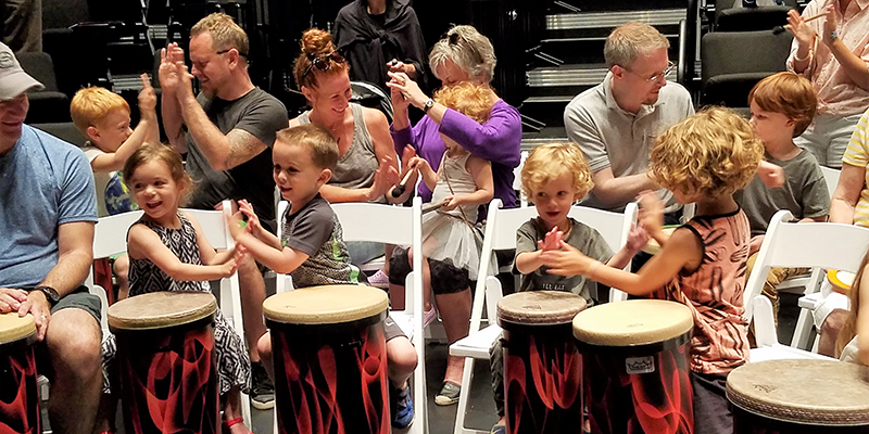 Young children play hand-clapping games with each other and the parents around them while sitting in foldable white chairs with bongo drums in the foreground.