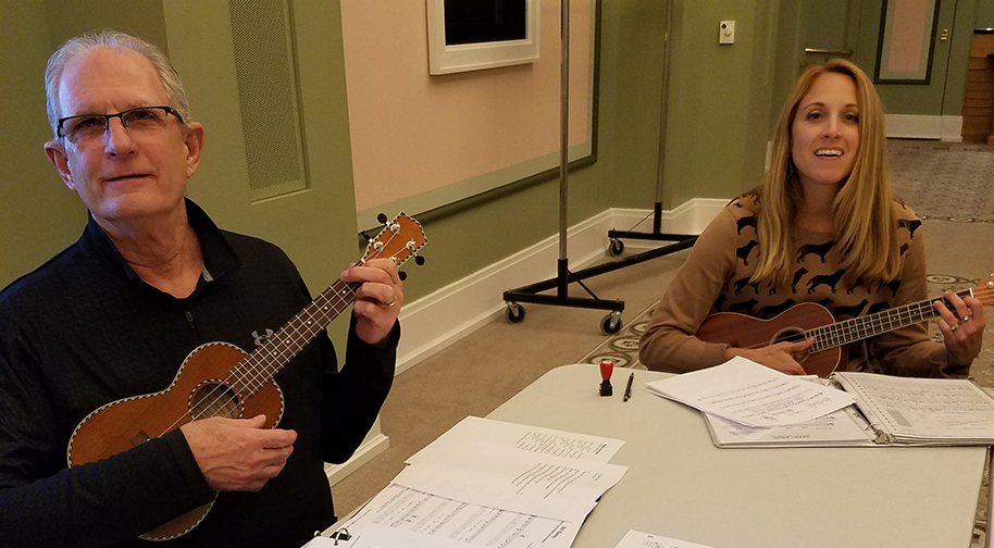 Two adult students strum their ukuleles at a folding plastic table with sheet music and lyrics in front of them.