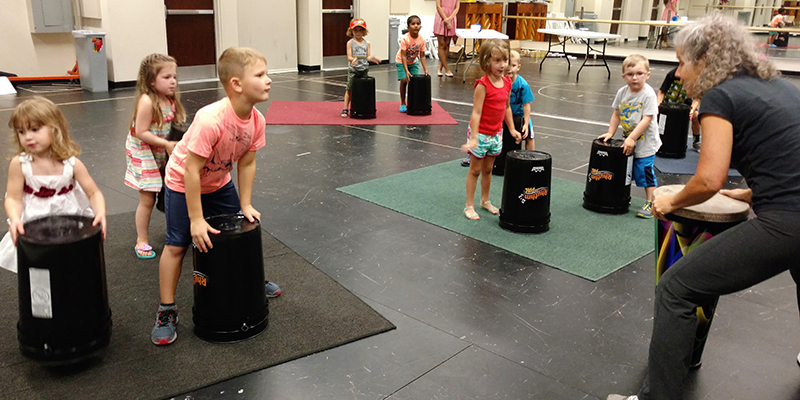 Children stand in front of overturned buckets, excitedly watching the instructor who is standing in front of them to demonstrate drumming on her hand drum.