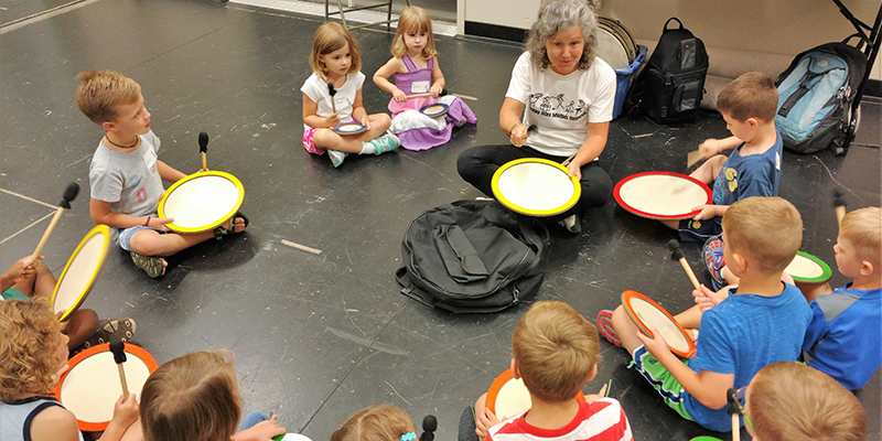 Children sit in a circle on a black dance floor with mallets and circular drumming instruments in their hands. They look toward the teacher, also sitting cross-legged on the floor with them, for instruction.