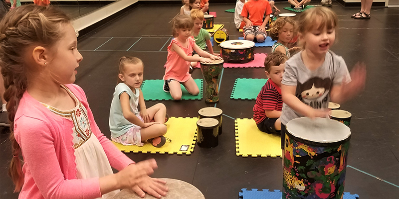 Children sit on colorful mats on the floor with various drums in front of them. Two children in particular are highlighted in the foreground, standing and drumming on their own instruments.