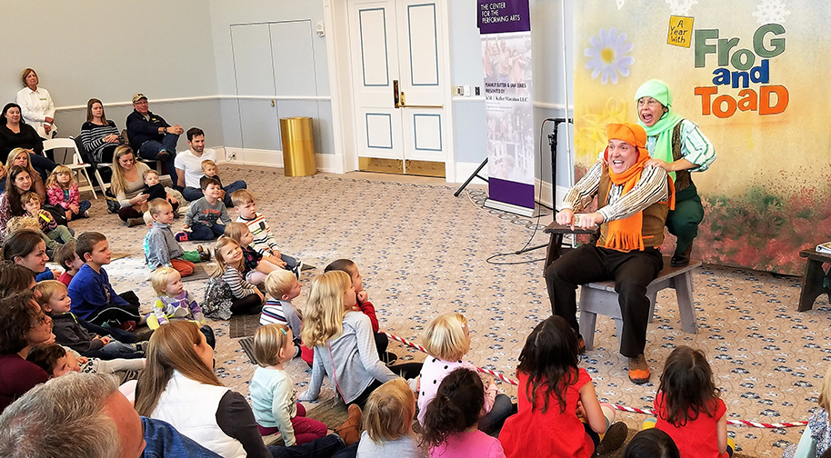 Children sit in rows on the floor watching two actors play "Frog and Toad". The actors are dressed in orange and green scarves, respectively, and one is holding on to the other as if they are on a high-speed vehicle together.