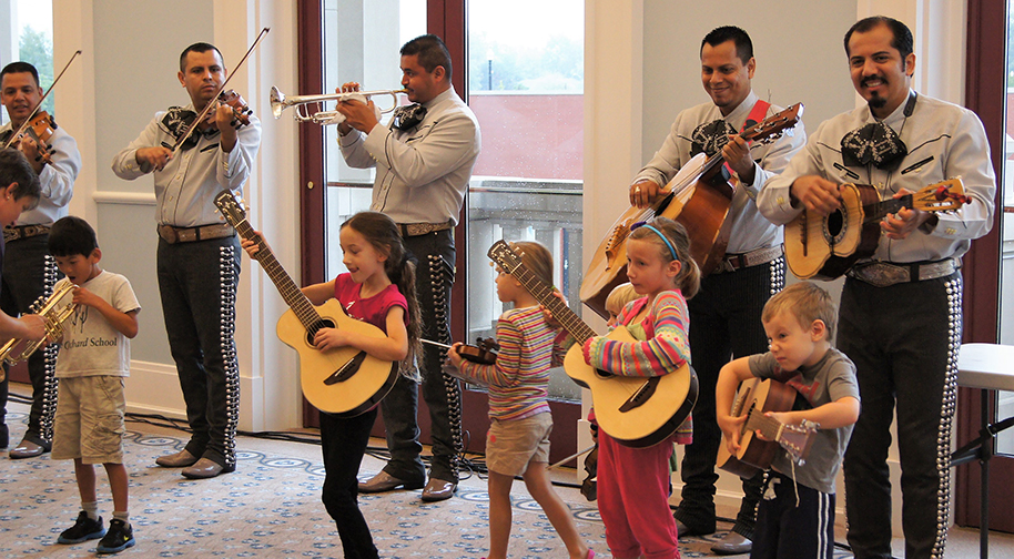 Five band members play various instruments (two violins, one silver trumpet, one guitar and one ukulele) in traditional mariachi garb. In front of them, young children try out their own instruments, including mini guitars and a trumpet.