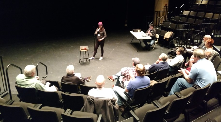 A young woman in a beanie holds a mic with a stool to her side on which sits a laptop. Other adults in the comedy class sit in chairs around her.