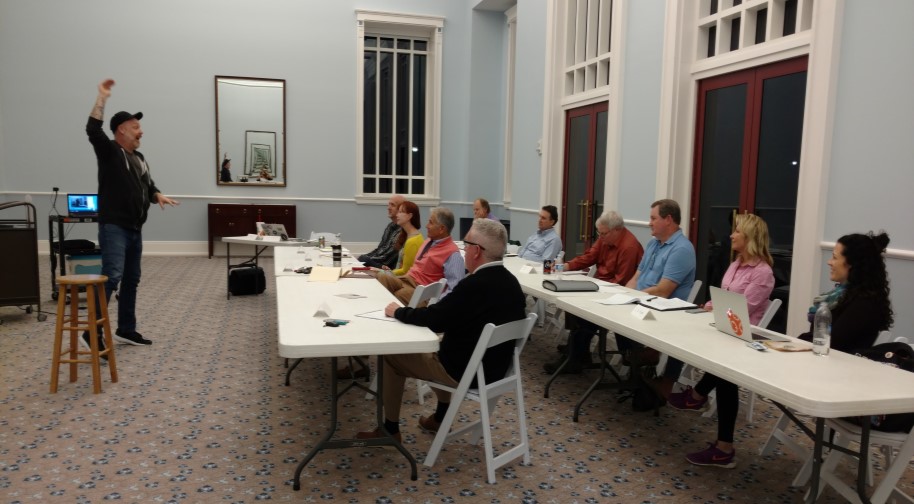 Adult students sit at two long plastic tables with notes in front of them. The instructor at the front of the class holds on hand in the air to emphasize a point.