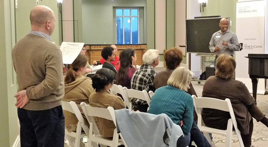 About a dozen people of varying ages sit in rows of folding white chairs while one man stands in front of the group and sings. An instructor stands in the back holding a piece of sheet music.