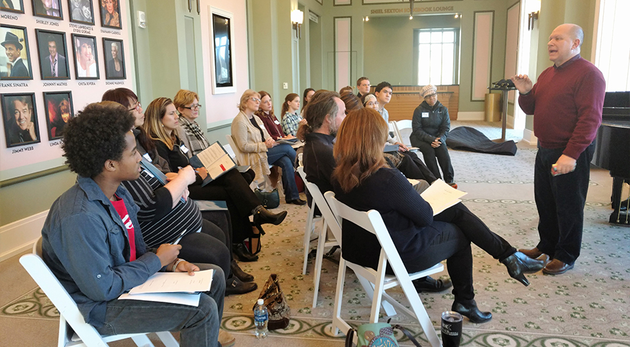 Almost twenty people of various ages sit in two rows of folding white chairs with folders and sheet music in their laps. They are watching an instruction speak or sing at the front of the class.