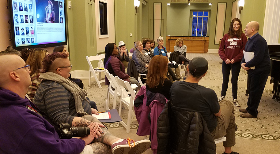 Almost twenty people of various ages sit in two rows of folding white chairs with folders and sheet music in their laps. An instructor stands at the front of the class giving instruction to one of the students.