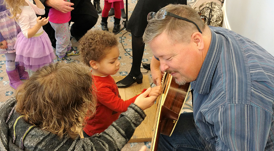 An adult gently takes the hand of a young child and strums it on the strings of a guitar being held by an adult in a striped blue shirt with spectacles on his head.