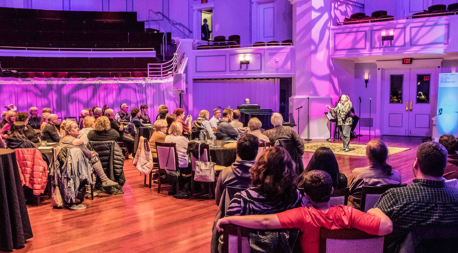 The Palladium is set with tables and chairs for a cabaret-style performance. People fill the seats and watch a woman singing into a microphone and extending her arm.