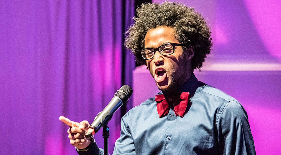 A young man in a blue button-up shirt and red bowtie sings into a microphone and emphasizes with his pointer finger.