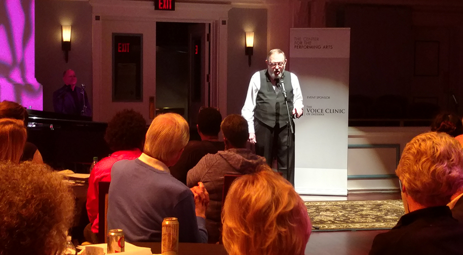 One man sings into a microphone in the spotlight. In the foreground are the backs of the audience's heads.