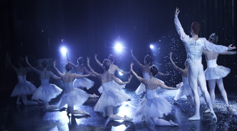 Rear view of a dozen actors onstage, dressed in white and raising their arms.