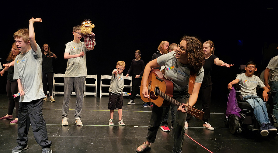 Prism Project campers dance in matching t-shirts alongside aides and a teacher playing an acoustic guitar.