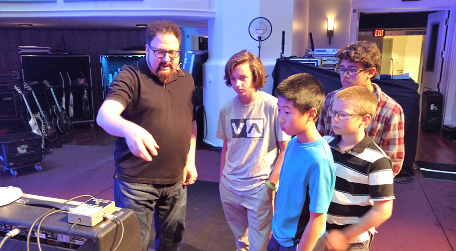 A man in a black shirt and black jeans points to technical theater equipment while four young boys look on.