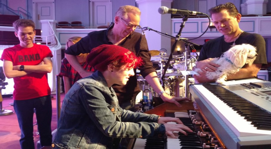 A young adult student in a red beanie and jean jacket plays a keyboard while several professional musicians look on. One is holding a small white dog.