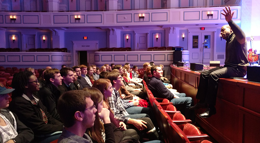 Young adult students sit in the third and fourth rows of the Palladium concert hall, listening intently to a musician onstage who is sharing about his career while holding his hand in the air to emphasize a point.