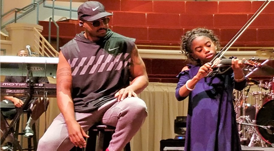 Damien Escobar, professional violinist, looks on as a young girl plays his violin. There is a drum set and a keyboard in the background.