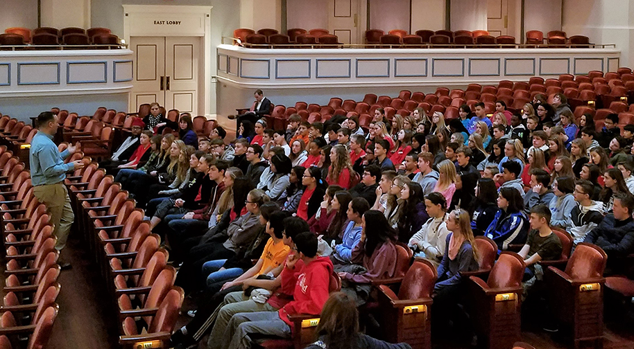 Nine rows in the Palladium are filled with young adult students who are watching and listening to an adult in the front who is wearing khakis and a blue button-up shirt.