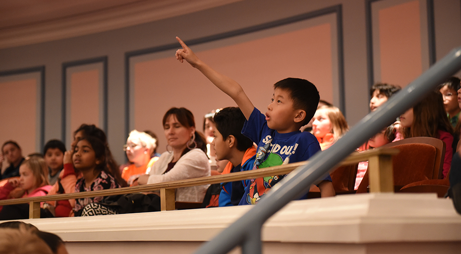 A young child excitedly points in the air from a balcony in the Palladium. Other adults and children sit in the seats around him.