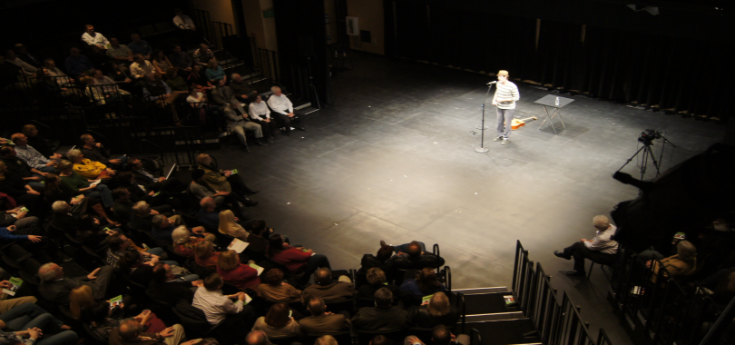 A person stands at a microphone surrounded by an audience in a black box theater.