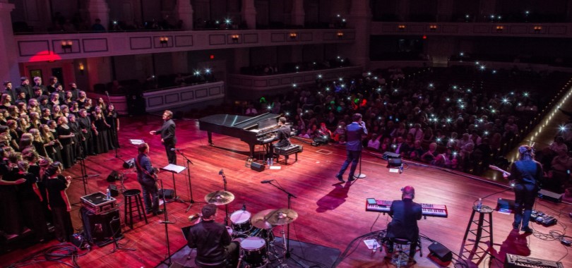 Viewed from behind, a band and choir perform onstage at the Palladium concert hall.