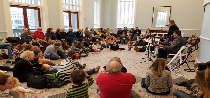 Families with small childen are seated on the floor of a large gathering space, watching musicians perform.