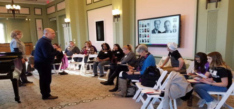 People are seated in white folding chairs listening to the instructor during a vocal class.