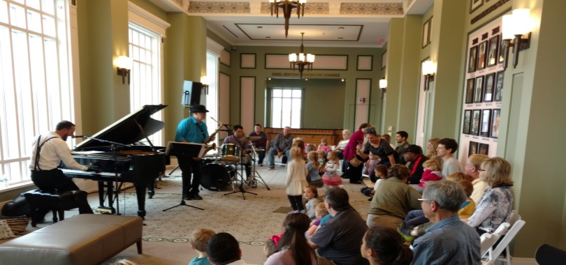 Families with small childen are seated in a carpeted lounge space watching musicians perform on piano, saxophone and drums.