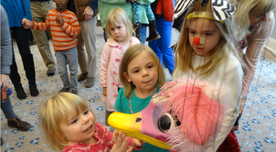 Young children excitedly crowd around a marionette shaped liked a duck with big pink feathers, a pink face and a yellow beak.