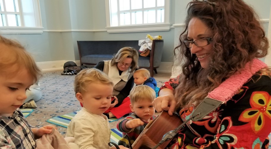 Three young children gather around a guitarist in a colorful floral shirt. They touch various parts of the guitar in fascination and wonder. In the background, a mom and her child inspect another instrument on the ground.