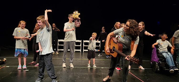 A woman with a guitar leads a group of kids in performing a song.