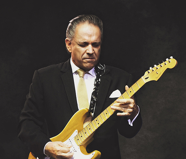 A man in a black suit plays a blonde Fender electric guitar