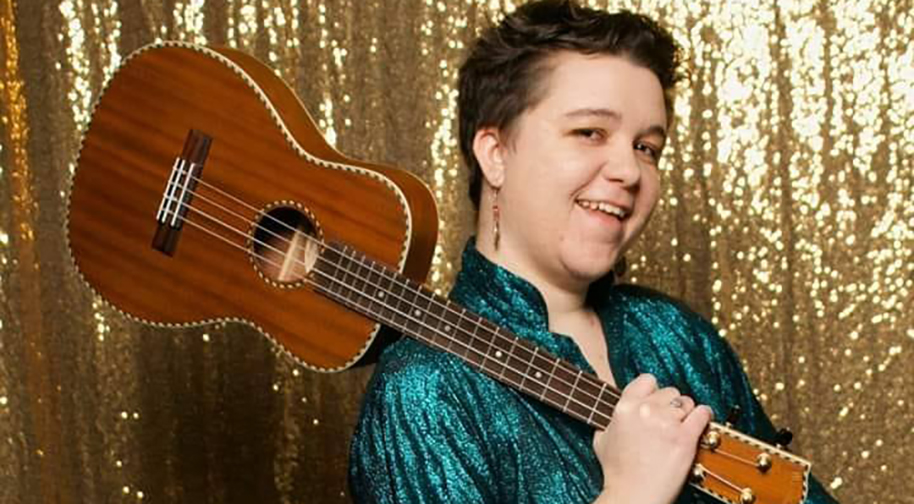 Meghan Martin, a white woman in her 20s, stands against a gold backdrop and wears an emerald green shirt. She holds a tenor ukulele over her shoulder.