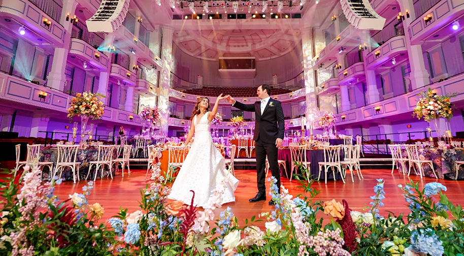 A bride and groom pose in front of floral arrangements on the Palladium stage.