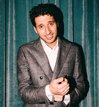 A man with dark curly hair wearing a suit stands in front of a blue curtain, hands clasped.