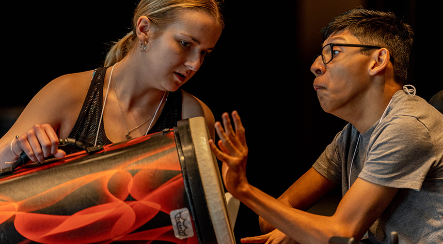 A Prism Project camper plays a conga-style drum being held by a teacher assistant.