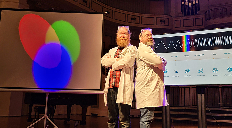 Two men in white lab coats and protective eyewear pose back to back in front of a screen displaying scientific information about light and sound wavelengths.