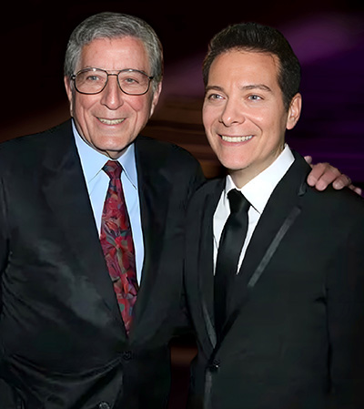 Singer Tony Bennett puts his arm around the shoulder of performer Michael Feinstein. Both men are wearing dark suits.