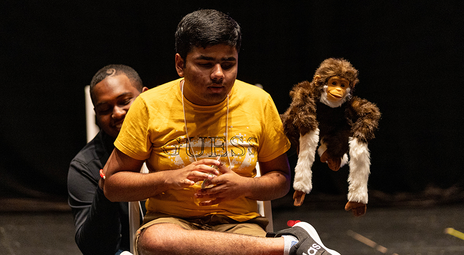 A Prism Project camper in a yellow t-shirt performs with the assistance of an aide holding a puppet.