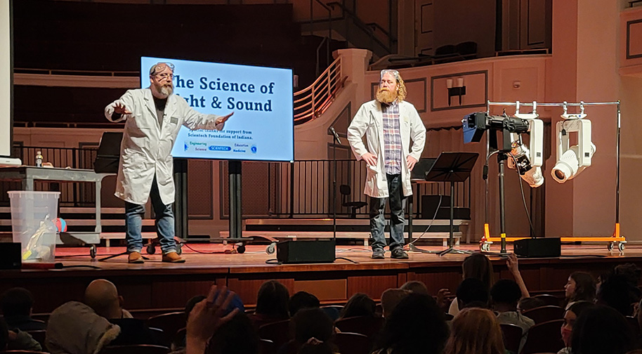 Two men in white lab coats and protective eyewear present in front of a screen reading The Science of Light & Sound.