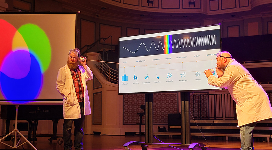 Two men in white lab coats and protective eyewear present in front of a screen displaying scientific information about light and sound wavelengths.