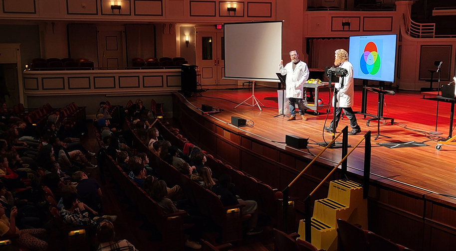 Two men in white lab coats and protective eyewear present in front of a screen displaying scientific information about light and sound wavelengths.