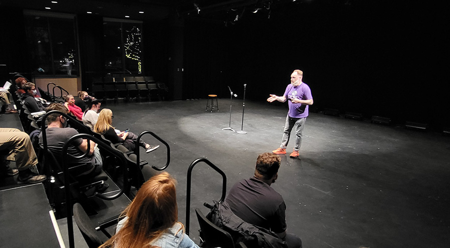 Professional comic Mat Alano-Martin stands on stage in the Studio Theater next to a microphone stand and a stool as he guides an Intro to Stand-Up Comedy class in forming their comedy set.