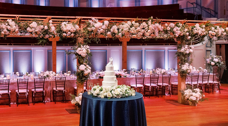 A pergola covered in white flowers and a tall white wedding cake all set on the Palladium stage.